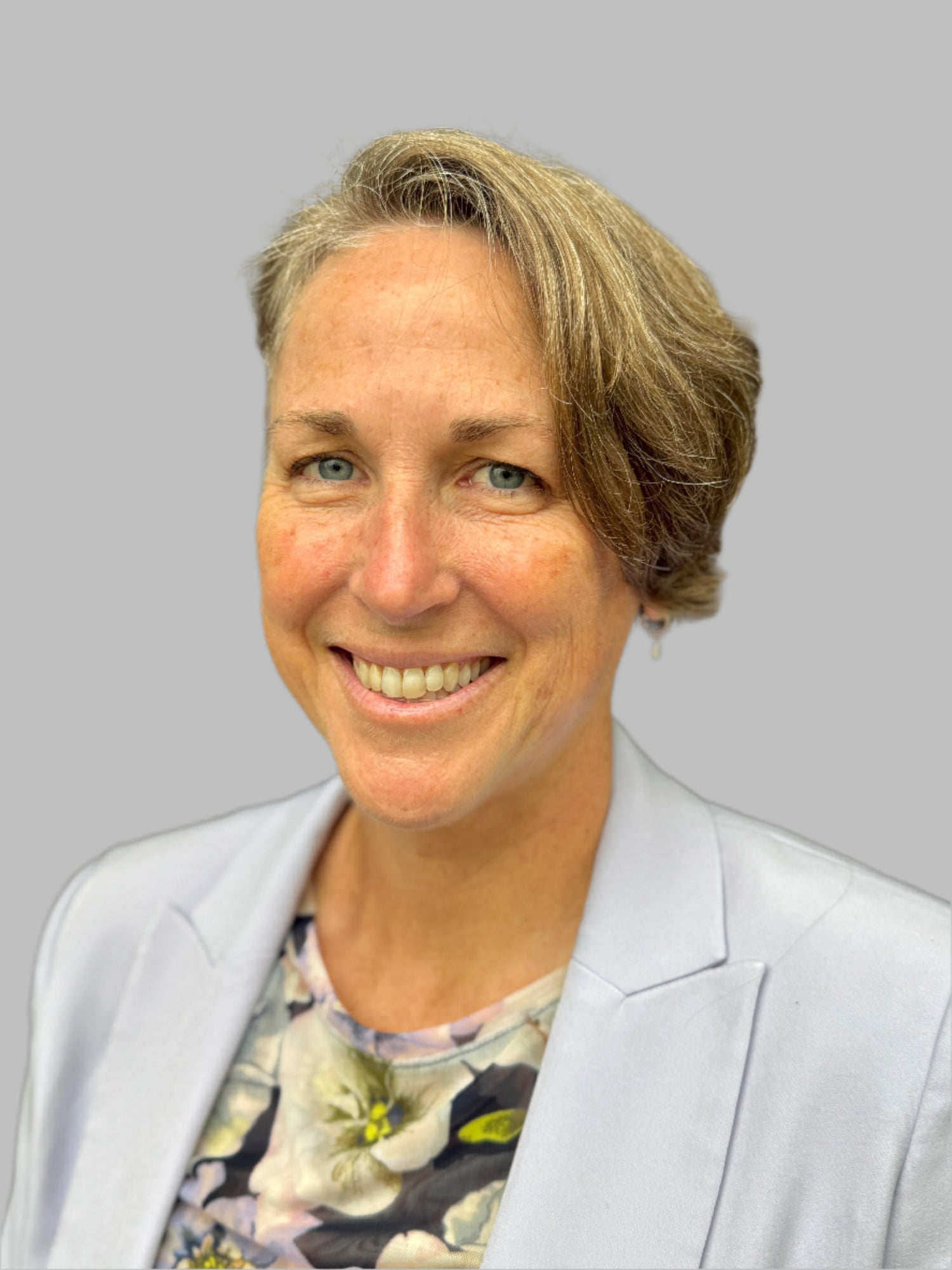 Suzen DuBre with short blonde hair wearing a light gray blazer over a floral shirt on a gray background.