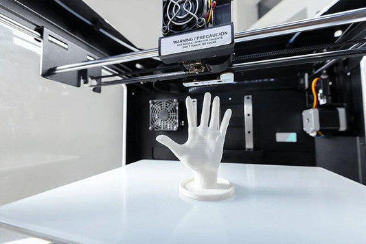A white 3D printer is printing a detailed model of a human hand, which stands upright on a circular base.